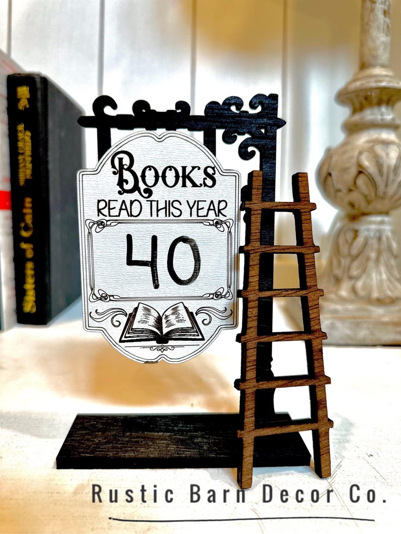 以下が含まれることがあります： 黒い梯子と「Books Read This Year 40」と書かれた白い看板が付いた木製看板。看板は黒い台座の上に置かれ、「Rustic Barn Decor Co.」の文字が書かれている