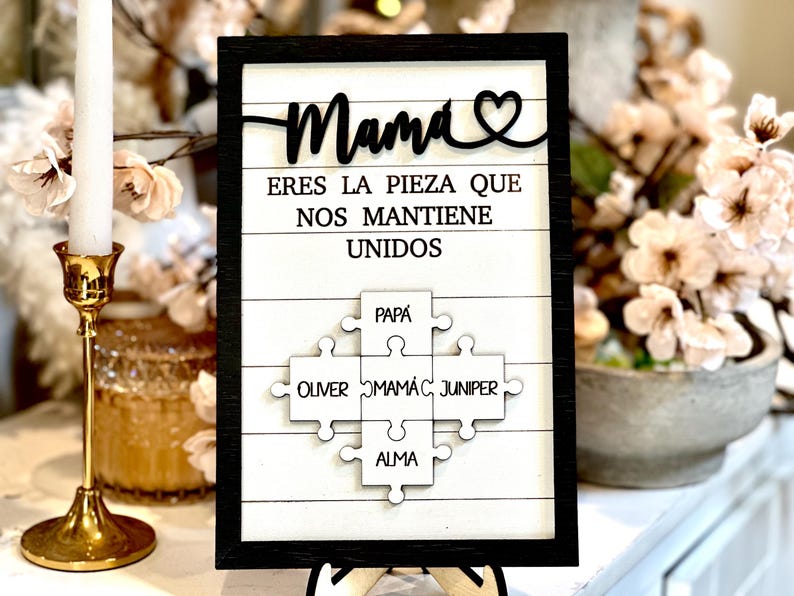 May include: A white wooden sign with black lettering that reads "Mama eres la pieza que nos mantiene unidos" with a heart symbol. The sign features a puzzle piece design with the names "Papa", "Oliver", "Mama", "Juniper", and "Alma" written on each piece.