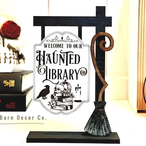 May include: A wooden sign with a black and white illustration of a haunted library. The sign is attached to a wooden post with a broom attached to the side. The sign reads "Welcome to our Haunted Library".