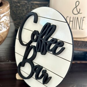 Pu&ograve; includere: Un cartello rotondo in legno bianco con lettere in legno dipinte di nero che compongono la scritta "Coffee Bar" su uno sfondo in legno sbiancato.