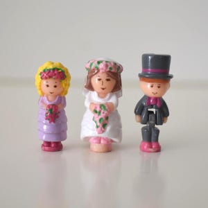 May include: Three small plastic figurines standing in a row. The left figure has yellow hair, a pink floral crown, and a purple dress. The center figure wears a white dress and a pink floral veil. The right figure wears a gray suit and top hat.