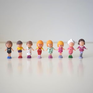 May include: A collection of eight small plastic dolls, each with a unique outfit and hairstyle. The dolls are holding hands, with a variety of colors in their clothing, including black, yellow, red, green, and pink. They stand on a white surface.