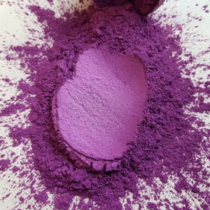 May include: Close-up of a pile of vibrant purple powder with a metallic sheen. The powder is scattered on a white surface, with a heart-shaped indentation in the center. The color is a deep, rich shade of violet.