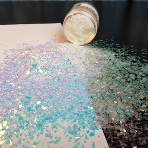 May include: A clear jar containing white iridescent glitter, spilling onto a white surface. The glitter is a mixture of small and large pieces, with a blue and purple iridescent sheen.