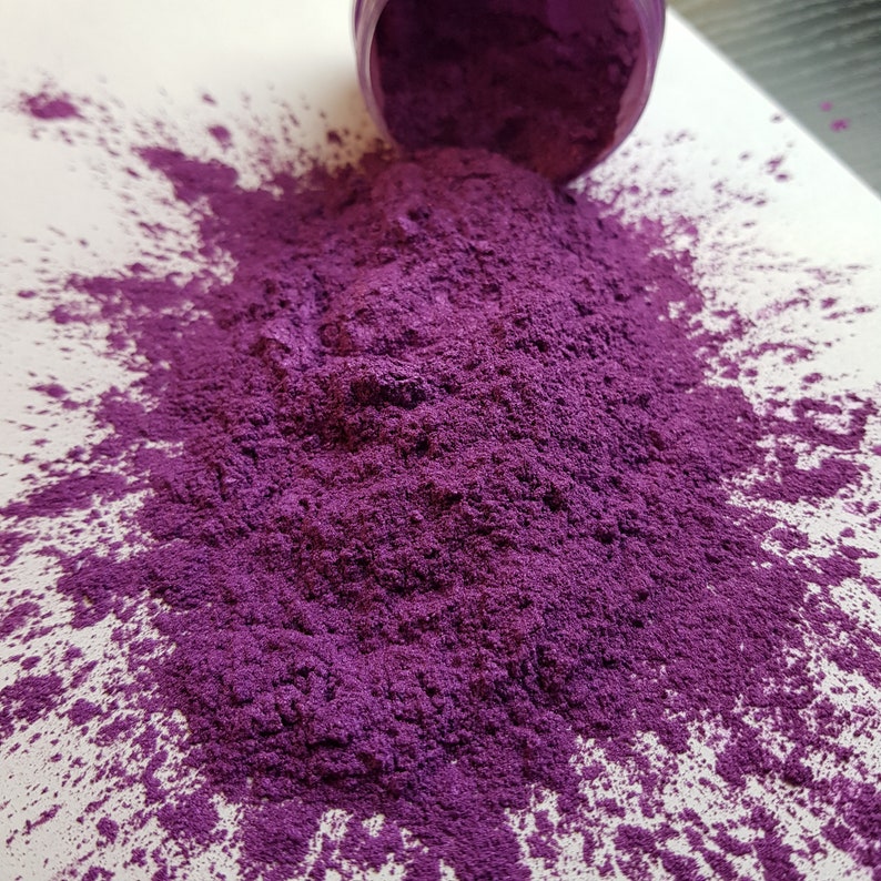 May include: A vibrant purple powder spills from a small container onto a white surface. The powder is finely ground and has a rich, saturated color. The container is a matching shade of purple.