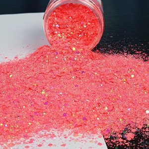 May include: A close-up of a jar of pink glitter with iridescent flakes. The glitter is spilling out of the jar onto a white surface.