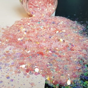 May include: A close-up of pink glitter spilling from a jar. The glitter is a mix of shapes and sizes, with iridescent and holographic elements. The text "Mystic Art Glitters" is visible.