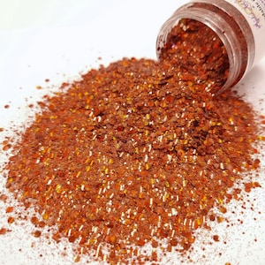 May include: Copper-coloured glitter spills from a clear jar. The glitter consists of small, irregularly shaped pieces that catch the light, creating a sparkling effect. The jar has a white label with purple text.