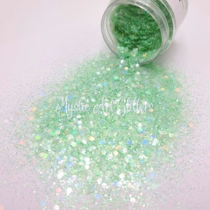 May include: A jar of green glitter with iridescent shimmer. The glitter is spilling out of the jar onto a white surface.
