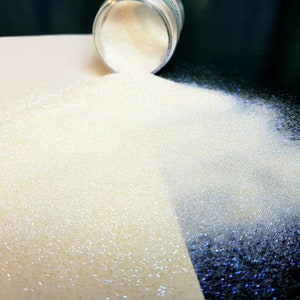 May include: Iridescent glitter spilling from a small clear jar. The glitter is a pale, creamy colour with a subtle shimmer. The glitter is scattered across a white and dark surface.