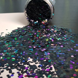 May include: A jar of iridescent black glitter with a rainbow sheen. The glitter is spilling out of the jar onto a white surface.
