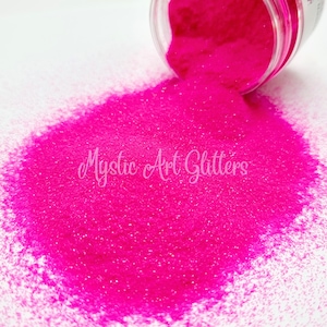 May include: A close-up of a bright pink glitter, with a small amount of glitter spilling out of a clear jar. The glitter is very fine and has a slight shimmer to it. The text "Mystic Art Glitters" is visible in the image.