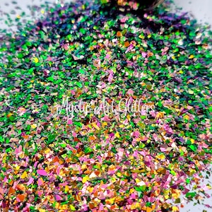 May include: A close-up of iridescent green, pink, and gold glitter flakes. The glitter is scattered on a white surface.
