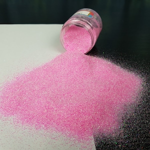 May include: Pink glitter with an iridescent shimmer is poured from a jar onto a white surface. The glitter is fine and has a subtle sparkle.