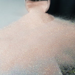 May include: A close-up of light pink glitter pouring from a clear container. The glitter is fine and sparkly, creating a shimmering effect. The background is a mix of black and white.