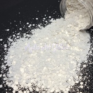 May include: White iridescent glitter in a jar, with some glitter spilling out onto a black surface. The glitter is a mix of small and large hexagons.