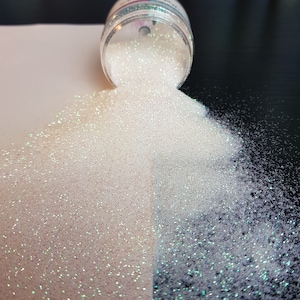May include: A clear jar of white glitter is tipped over, spilling onto a white and black surface. The glitter is a fine powder with iridescent flecks, creating a sparkling effect. The glitter is a craft supply.