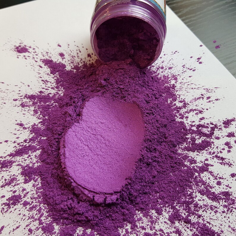 May include: A spilled container of purple pigment powder on a white surface. The powder forms a heart shape in the center, with a lighter shade of purple. The container is partially visible, with a purple lid.
