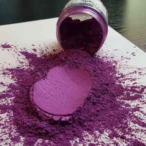 May include: A jar of purple pigment powder is tipped over, spilling onto a white surface. The powder forms a heart shape with a surrounding cloud of scattered pigment. The jar's label is partially visible.