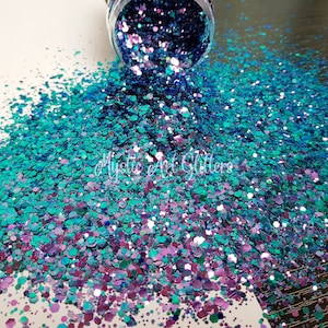 May include: A close-up of a pile of iridescent glitter in shades of teal, purple, and blue, spilling from a clear container. The glitter is scattered across a white surface. The text "Mystic Art Glitters" is visible.