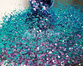 May include: A close-up of a pile of iridescent glitter in shades of teal, purple, and blue, spilling from a clear container. The glitter is scattered across a white surface. The text "Mystic Art Glitters" is visible.