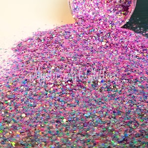 May include: A close-up of iridescent pink, purple, and blue glitter. The glitter is scattered on a white surface.