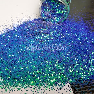 May include: A close-up of a pile of iridescent blue and green glitter. The glitter is scattered on a white surface and has a holographic finish.
