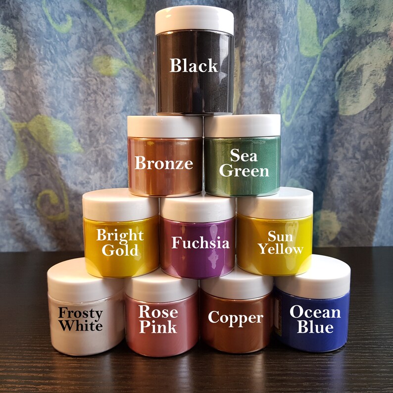 May include: A collection of ten jars of colored pigment powders with white lids. Colors include black, bronze, sea green, bright gold, fuchsia, sun yellow, frosty white, rose pink, copper, and ocean blue. Each jar has the color name printed on it.