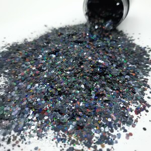 May include: A pile of black, silver, and iridescent glitter with a rainbow sheen. The glitter is loose and scattered on a white surface.