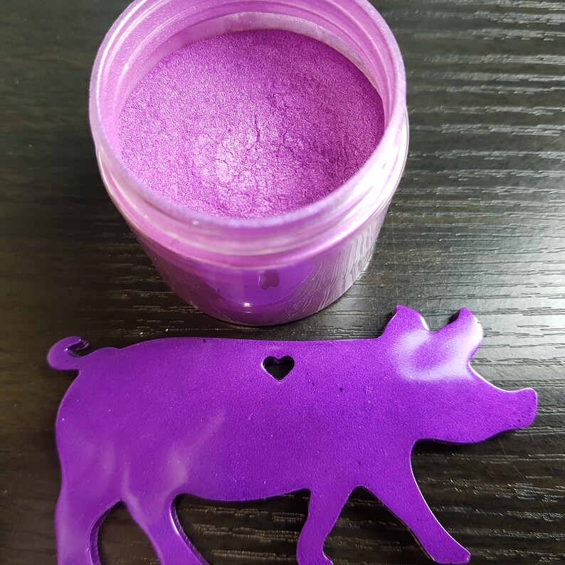 May include: A jar of vibrant purple powder sits next to a purple pig-shaped ornament with a heart cutout. The powder has a shimmering, metallic finish. The items are on a dark wood surface.