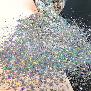 May include: A close-up of iridescent glitter in a variety of shapes and sizes. The glitter is a mix of silver, blue, green, and rainbow colours. The glitter is spilling out of a jar onto a white surface.