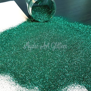 May include: A close-up of emerald green glitter scattered from a clear jar. The glitter particles are small and shiny. The image includes the text "Mystic Art Glitters".