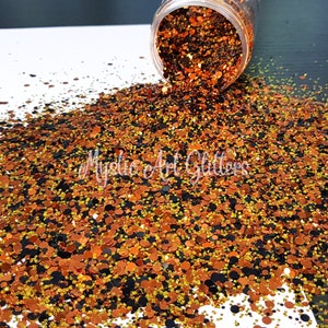 May include: A close-up of a jar of orange, gold, and black glitter. The glitter is spilling out of the jar and onto a white surface. The glitter is in various sizes and shapes.