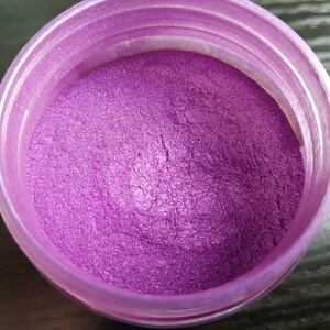 May include: A container filled with vibrant, shimmering purple powder. The powder has a metallic sheen, reflecting light. The container is a translucent, light purple color. The product appears to be a cosmetic pigment or craft supply.