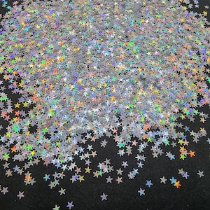 May include: A close-up of a pile of silver star-shaped glitter. The glitter has a holographic effect, reflecting a rainbow of colours. The stars are scattered across a black surface, creating a sparkling effect.