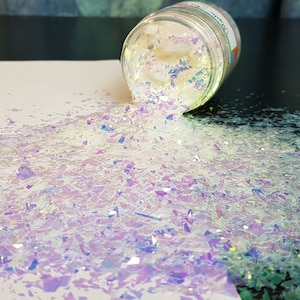 May include: A clear glass jar filled with iridescent white glitter, spilling out onto a white surface. The glitter is made up of small, irregular pieces that catch the light and sparkle.