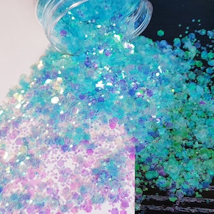 May include: A close-up of iridescent blue and purple glitter, with some of the glitter spilling out of a clear jar onto a white surface. The glitter is a mix of small and large hexagons and circles.