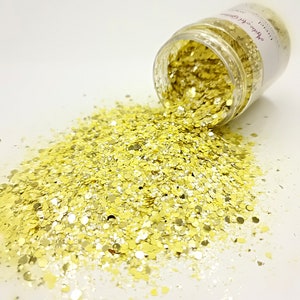 May include: A clear plastic jar of gold glitter is tipped over, spilling onto a white surface. The glitter is a mix of round and irregular shapes, reflecting light. The jar has a label with text.