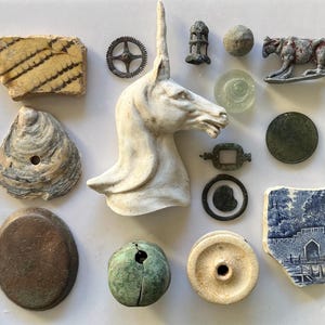 Może przedstawiać: Kolekcja różnorodnych artefaktów, w tym białe popiersie jednorożca, fragment wzorzystej ceramiki, przewiercona muszla ostrygi, metalowe koło zębate, mała figurka zwierzęcia, niebiesko-biały odłamek ceramiki, zielona kula oraz różne metalowe żetony.
