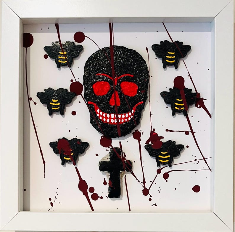 May include: A framed wall art piece featuring a textured black skull with red eye sockets and a white-toothed grin, surrounded by six small black and yellow bees and a black cross, all set against a white background with red paint splatters.