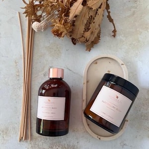 May include: A brown glass diffuser bottle with a gold lid and a label that reads "Australian Bush" and a brown glass candle jar with a black lid and a label that reads "Australian Bush". Both bottles are on a white oval tray with a textured surface.