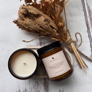 May include: Two brown glass candle jars with lids. The jar on the right has a label that reads "AshWood & Co. Handmade Soy Candles" and "Vanilla, Patchouli + Sandalwood" with "Sydney, Australia" at the bottom. The jar on the left is filled with a white candle.