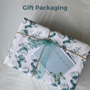Op de afbeelding: Een witte cadeaudoos verpakt in papier met een patroon van blauwe en groene eucalyptusbladeren. De doos is vastgebonden met touw en een blauw lint. Een klein blauw kaartje met de tekst "Thinking of you" is aan de doos bevestigd.