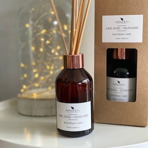 May include: A brown glass bottle with a copper coloured lid and a label that reads "AskWood & Co Lime, Basil, Mandarin Reed Diffuser | 140ml". The bottle is sitting on a white surface with a wicker basket underneath. The bottle is surrounded by wooden sticks.