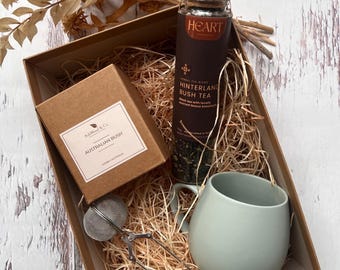 Op de afbeelding: Een bruine cadeaudoos gevuld met gescheurd papier bevat een mok, een pot met losse thee met het label "Hinterland Bush Tea", een kaars met het label "Australian Bush" en een theezeef.