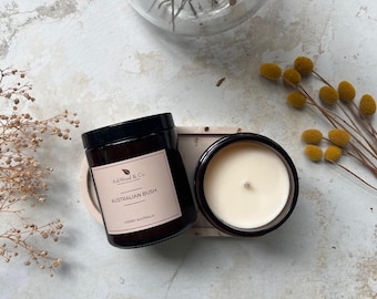Op de afbeelding: Twee bruine glazen kaarsenpotten met zwarte deksels. De kaars links heeft een label met de tekst "AshWood & Co. Handmade Soy Candles" en "AUSTRALIAN BUSH" met de tekst "Sydney Australia" eronder. De kaars rechts is aangestoken en heeft een wit wasoppervlak.