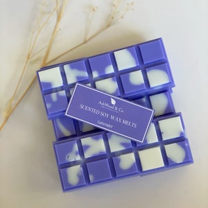 Op de afbeelding: Een stapel paarse en witte geurende sojawas smelt in een rasterpatroon. Het label luidt "AshWood & Co. Scented Soy Wax Melts Lavender".