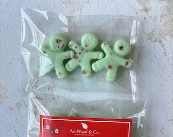 Op de afbeelding: Drie groene gemberman-vormige sojawas smelt in een doorzichtige plastic zak met een rood label waarop staat "AshWood & Co. Hand Poured Soy Candles" en "Scented Soy Wax Melts Santa's Gingerbread".