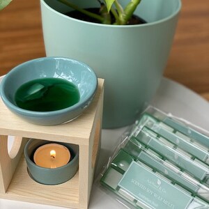 Op de afbeelding: Een houten wax warmer met een teal keramische kom gevuld met groene wax melts. Een teal theelichtje zit in de warmer. Een verpakking met groene en witte geurende sojawax melts ligt op een wit oppervlak. De verpakking heeft de tekst "Scented Soy Wax Melts Australian Made".