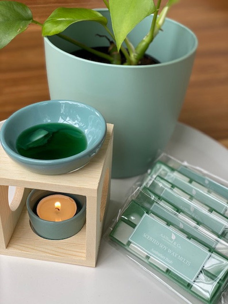 Op de afbeelding: Een houten wax warmer met een teal keramische kom gevuld met groene wax melts. Een teal theelichtje zit in de warmer. Een verpakking met groene en witte geurende sojawax melts ligt op een wit oppervlak. De verpakking heeft de tekst "Scented Soy Wax Melts Australian Made".